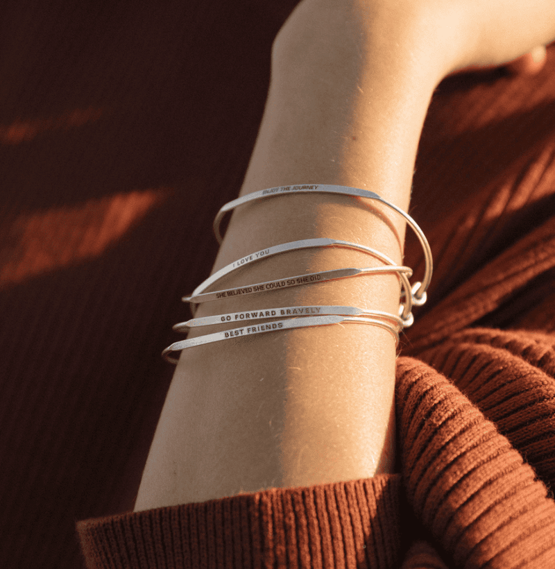 Stack of silver bangle bracelets with words engraved on them. Words include "THE WORLD IS YOURS". 
