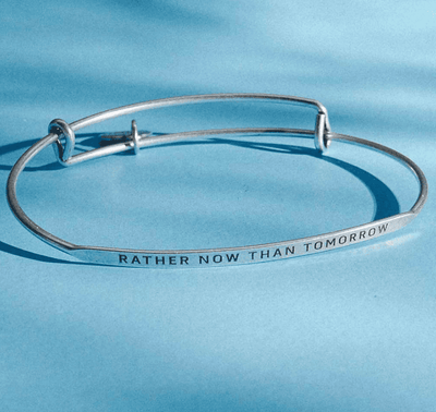 Silver bangle bracelet with the words "Rather Now Than Tomorrow" engraved on it. 
