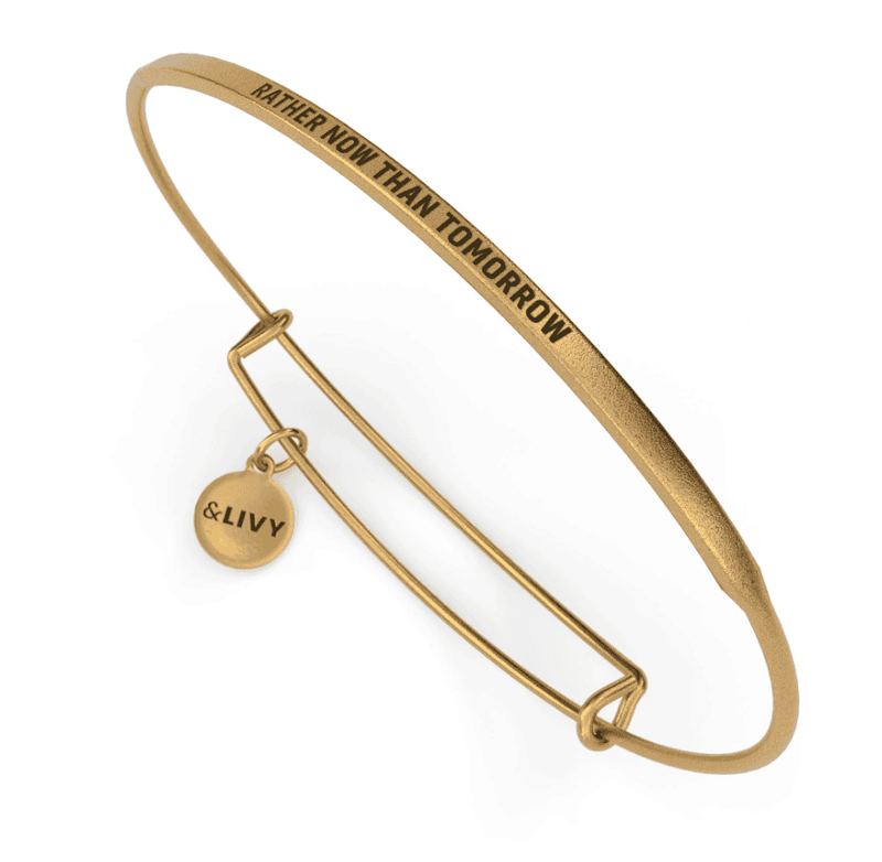 Gold bangle bracelet with the words "Rather Now Than Tomorrow" engraved on it. 