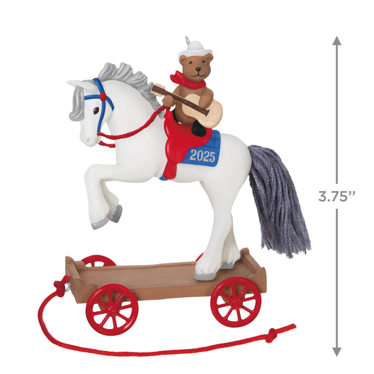 2025 ornament of a teddy bear cowboy on a white pony with red wheels, wearing a hat and scarf, strumming a guitar on a rolling wood-tone base.