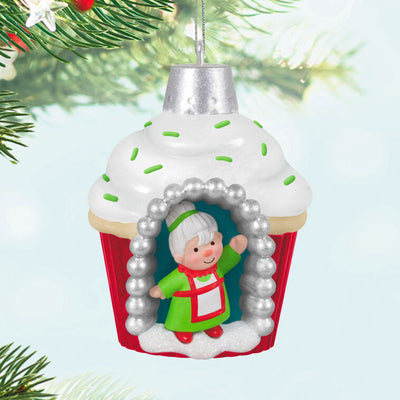Mini cupcake-shaped ornament with Mrs. Claus waving from a silver-dotted doorway, topped with white icing and green sprinkles.