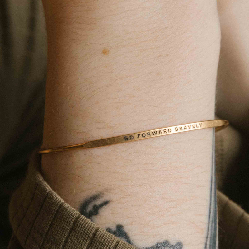 Gold bangle bracelet with the words "go forward bravely" engraved on it.