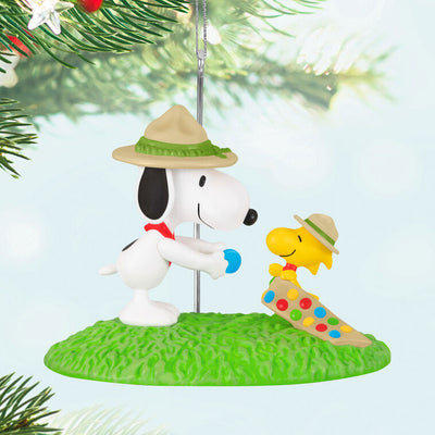 Snoopy in scout hat presenting merit badge to Woodstock in sash, standing on green grass, Hallmark Peanuts Christmas ornament collectible
