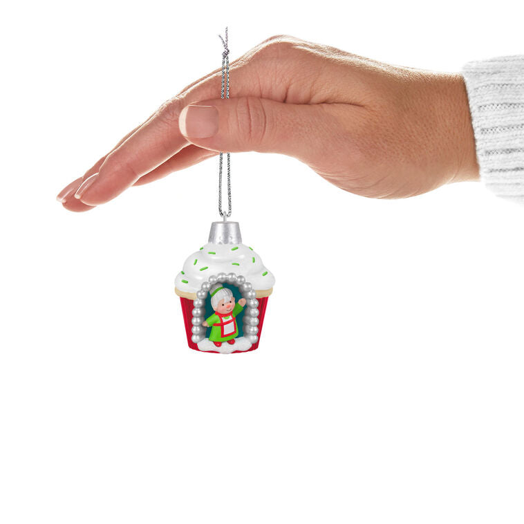 Mini cupcake-shaped ornament with Mrs. Claus waving from a silver-dotted doorway, topped with white icing and green sprinkles.