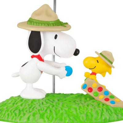 Snoopy in scout hat presenting merit badge to Woodstock in sash, standing on green grass, Hallmark Peanuts Christmas ornament collectible

