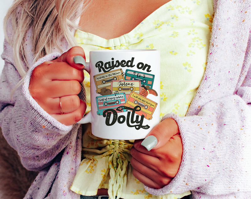 White 11oz coffee mug with pink “Raised on Dolly” country music design. Double-sided print, ceramic mug.

