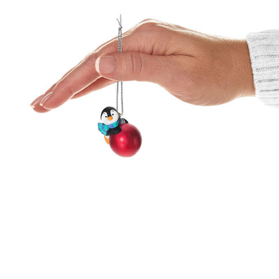 Mini penguin ornament holding a large red Christmas ball, wearing a blue plaid scarf with cheerful expression — Petite Penguins series.