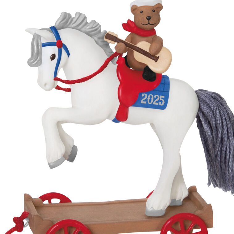 2025 ornament of a teddy bear cowboy on a white pony with red wheels, wearing a hat and scarf, strumming a guitar on a rolling wood-tone base.