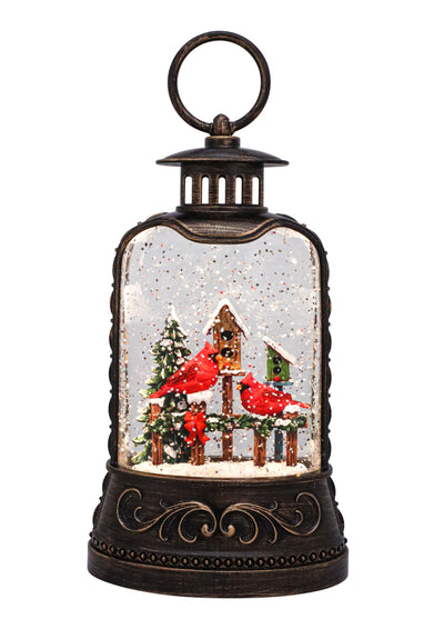 Bronze lantern with red cardinal on snowy birdhouse, swirling glitter, and warm LED glow.
