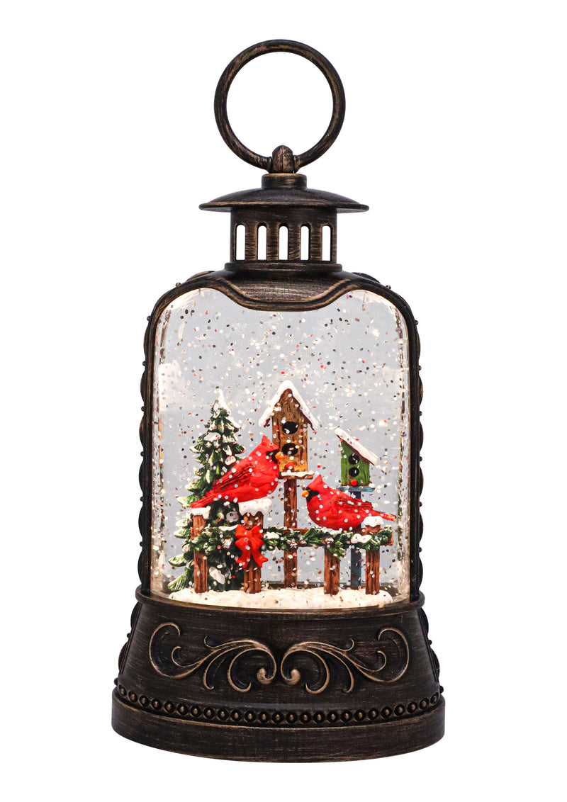 Bronze lantern with red cardinal on snowy birdhouse, swirling glitter, and warm LED glow.
