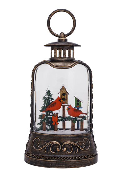 Bronze lantern with red cardinal on snowy birdhouse, swirling glitter, and warm LED glow.
