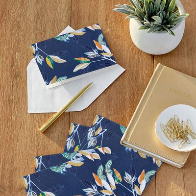 Elegant blank note cards with gold-accented tropical leaves on navy background, boxed set of 10 with white envelopes.
