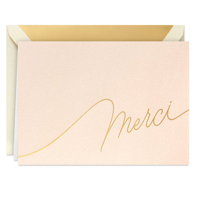 Blush pink thank-you cards with gold foil “Merci” script lettering and matching foil-lined envelopes in a white display box.
