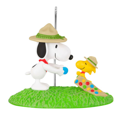 Snoopy in scout hat presenting merit badge to Woodstock in sash, standing on green grass, Hallmark Peanuts Christmas ornament collectible