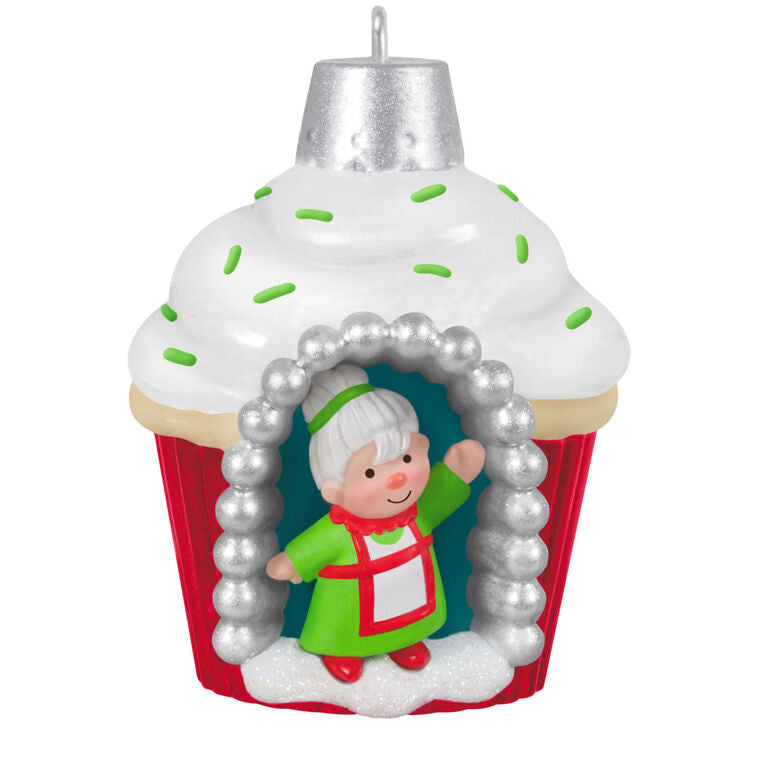 Mini cupcake-shaped ornament with Mrs. Claus waving from a silver-dotted doorway, topped with white icing and green sprinkles.