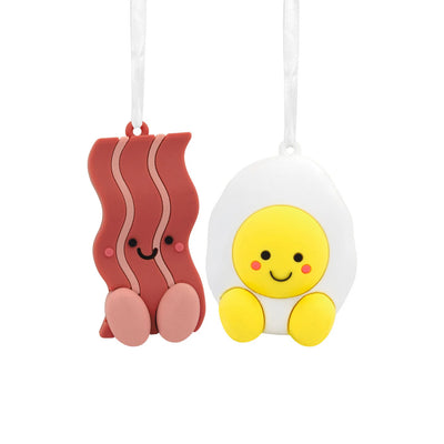 Christmas ornament set featuring two rubber ornaments: a smiling bacon slice and a smiling fried egg. They are connected by a red ribbon.