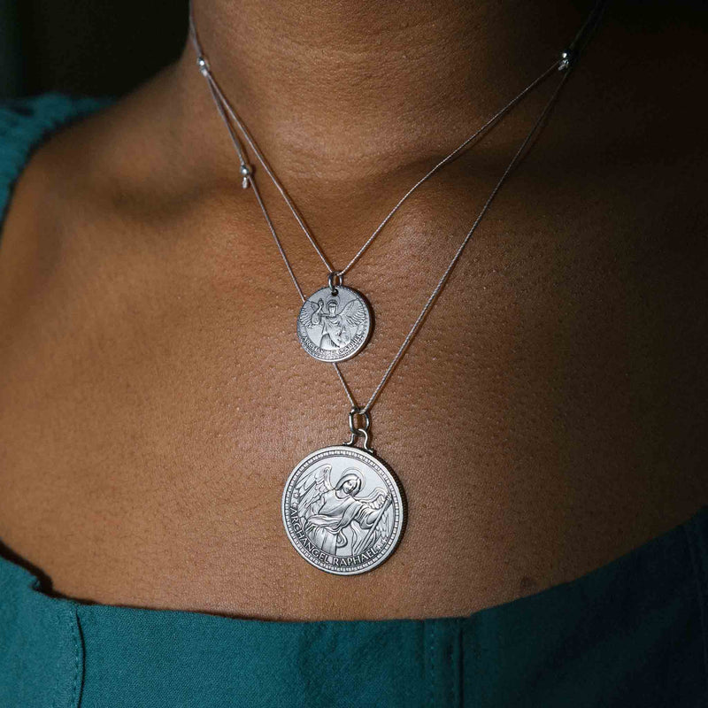 a silver necklace with two religious medallions. The medallions are round and  text “ARCHANGEL RAPHAEL.” is written in a loop around the rim of the medallion.