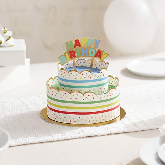 Hallmark 3D pop-up birthday cake paper decor with rainbow stripes, sprinkles, gold foil accents, and "Happy Birthday" banner.

