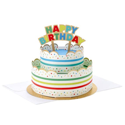 Hallmark 3D pop-up birthday cake paper decor with rainbow stripes, sprinkles, gold foil accents, and "Happy Birthday" banner.

