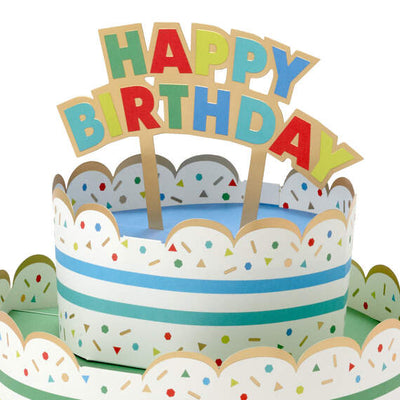 Hallmark 3D pop-up birthday cake paper decor with rainbow stripes, sprinkles, gold foil accents, and "Happy Birthday" banner.

