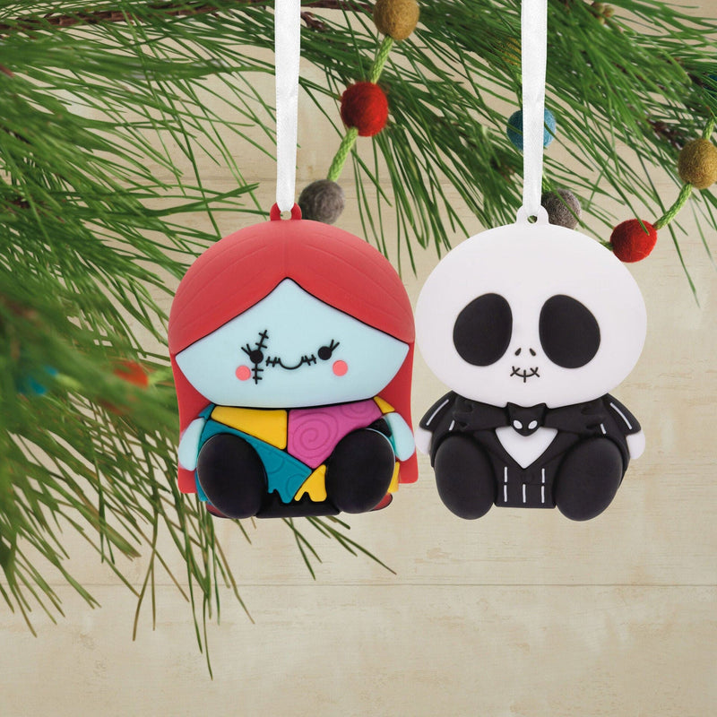Two Nightmare Before Christmas ornaments featuring Jack Skellington and Sally. Jack is a skeleton with a pinstriped suit and bat bowtie. Sally has ragdoll stitching and a patchwork dress. They are connected by a red ribbon
