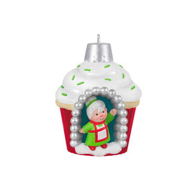 Mini cupcake-shaped ornament with Mrs. Claus waving from a silver-dotted doorway, topped with white icing and green sprinkles.