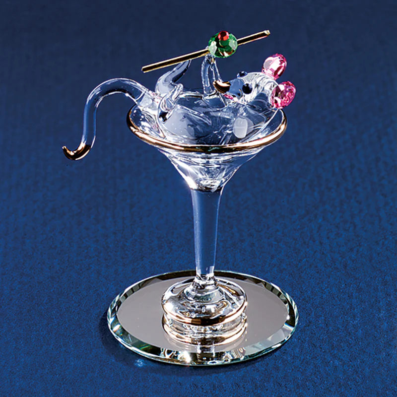 Handcrafted glass mouse holding olive, sitting in martini glass on beveled mirror.
