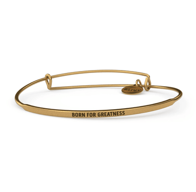 Gold bangle bracelet with the words "BORN FOR GREATNESS" engraved on it. The lettering is cursive.