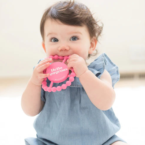 Pink round silicone teether with beaded edges and "Hello Gorgeous" phrase, designed for babies to chew and grip easily.