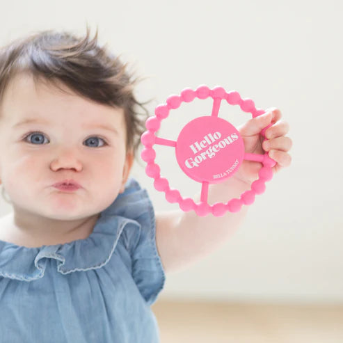 Pink round silicone teether with beaded edges and "Hello Gorgeous" phrase, designed for babies to chew and grip easily.