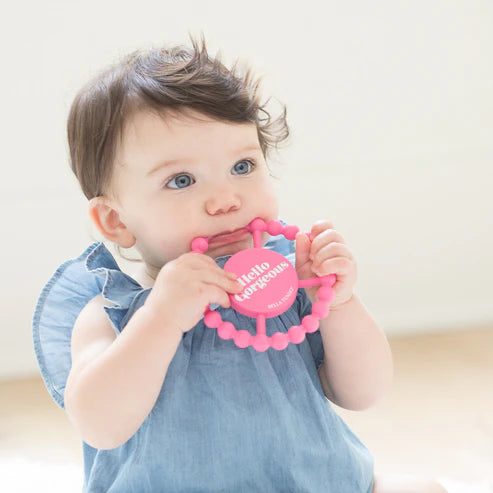 Pink round silicone teether with beaded edges and "Hello Gorgeous" phrase, designed for babies to chew and grip easily.