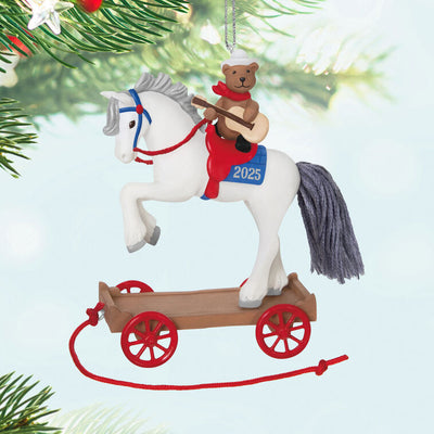 2025 ornament of a teddy bear cowboy on a white pony with red wheels, wearing a hat and scarf, strumming a guitar on a rolling wood-tone base.