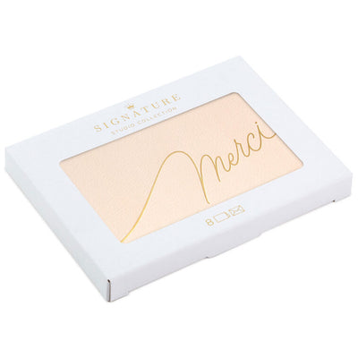 Blush pink thank-you cards with gold foil “Merci” script lettering and matching foil-lined envelopes in a white display box.
