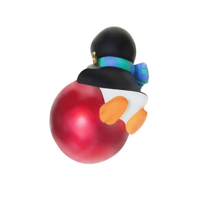 Mini penguin ornament holding a large red Christmas ball, wearing a blue plaid scarf with cheerful expression — Petite Penguins series.