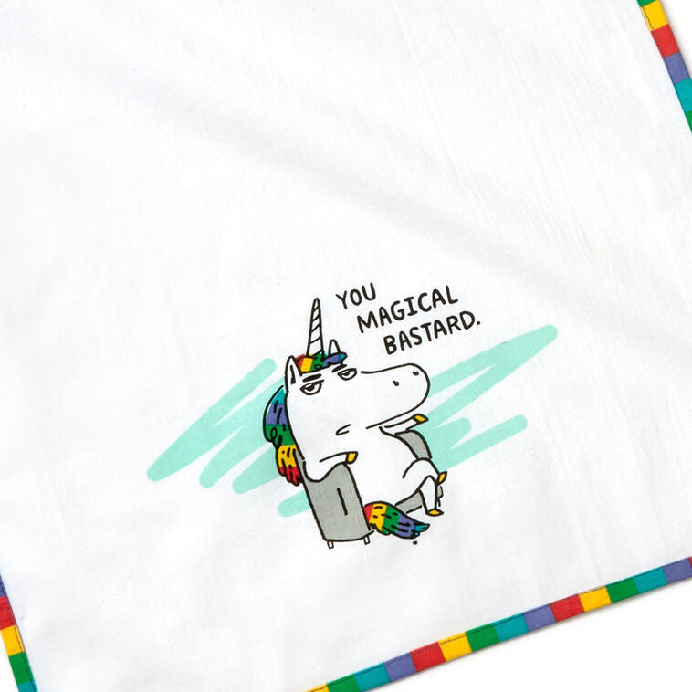 White cotton tea towel with rainbow-striped border and unicorn in chair, featuring the message: “You magical bastard.”