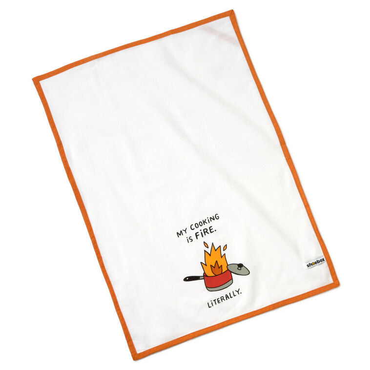 White cotton tea towel with orange border, featuring a flaming pot illustration and the message: “My cooking is fire.