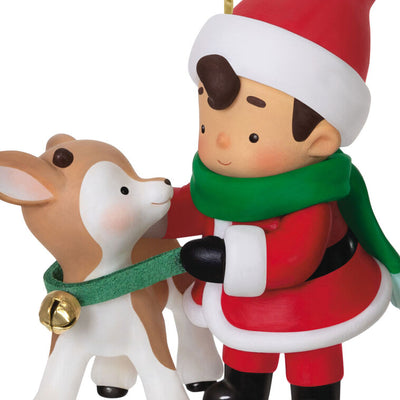 Hallmark Keepsake Young Santa ornament—Santa as a boy in red hat and coat, green scarf, placing green collar with gold bell on young reindeer
