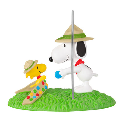 Snoopy in scout hat presenting merit badge to Woodstock in sash, standing on green grass, Hallmark Peanuts Christmas ornament collectible
