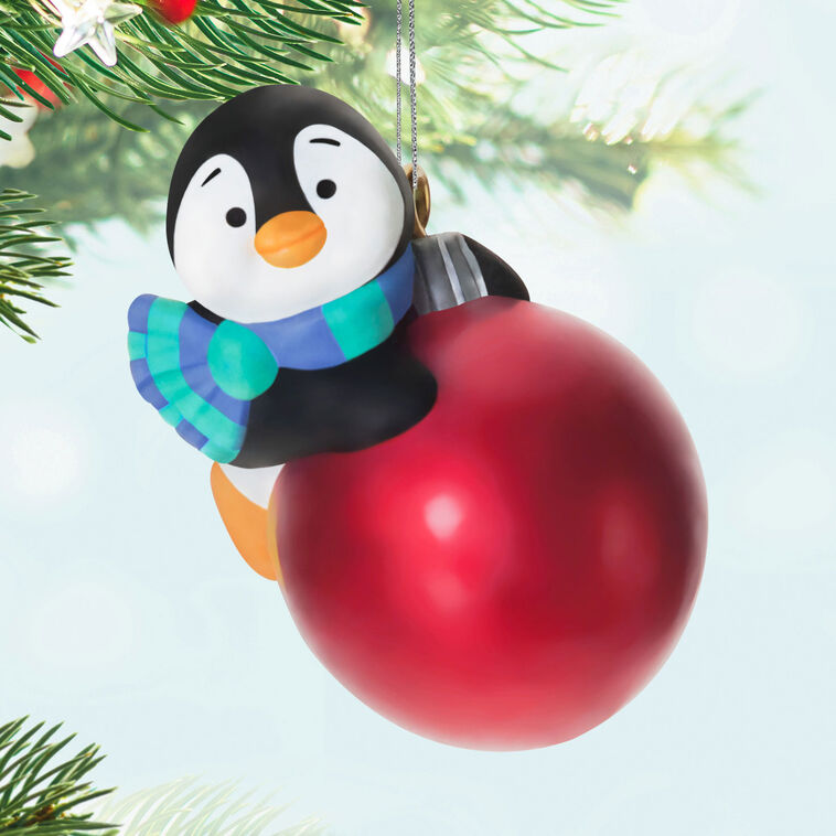 Mini penguin ornament holding a large red Christmas ball, wearing a blue plaid scarf with cheerful expression — Petite Penguins series.