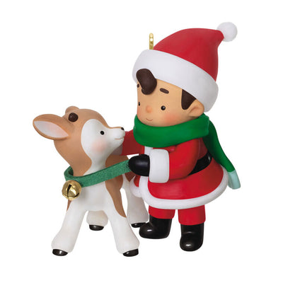 Hallmark Keepsake Young Santa ornament—Santa as a boy in red hat and coat, green scarf, placing green collar with gold bell on young reindeer
