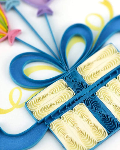 Quilled birthday card with three colorful balloons tied to a gift box with a blue bow and “Happy Birthday” message—playful and festive design.