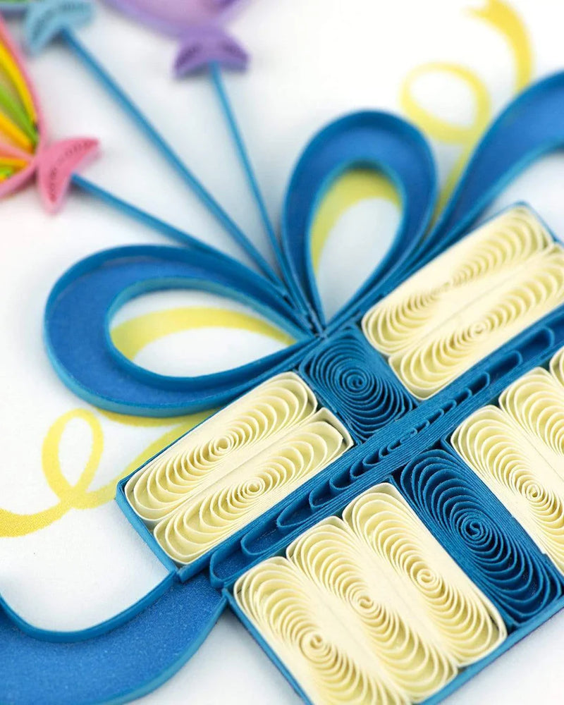 Quilled birthday card with three colorful balloons tied to a gift box with a blue bow and “Happy Birthday” message—playful and festive design.