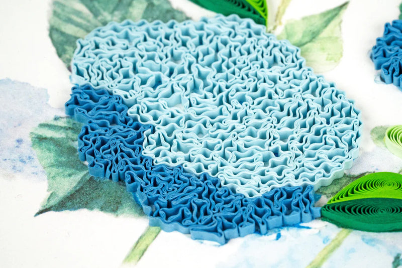 Quilled card showcasing a cluster of vibrant blue hydrangeas with green leaves, delicately handcrafted on a soft white background.