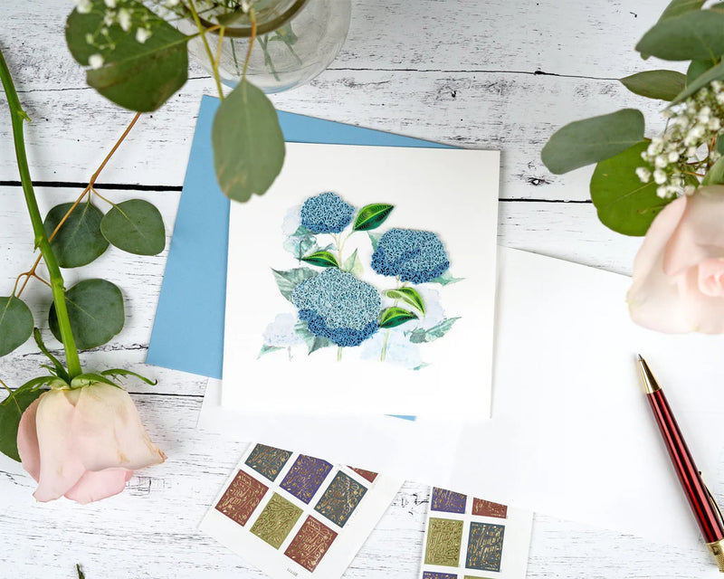 Quilled card showcasing a cluster of vibrant blue hydrangeas with green leaves, delicately handcrafted on a soft white background.