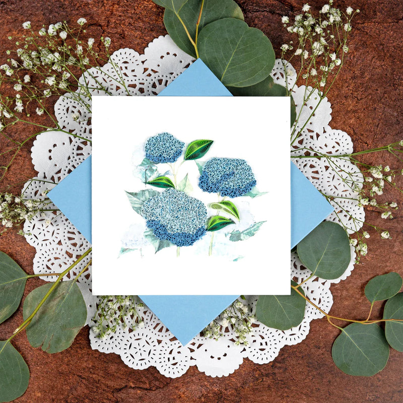 Quilled card showcasing a cluster of vibrant blue hydrangeas with green leaves, delicately handcrafted on a soft white background.