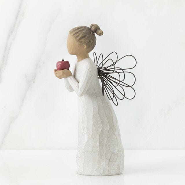 The figurine features an angel with outstretched arms clutching a bright red apple