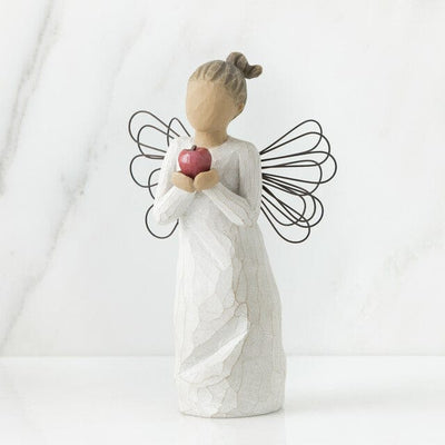 The figurine features an angel with outstretched arms clutching a bright red apple